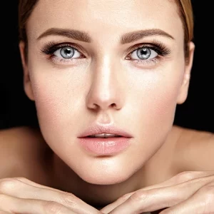 Close-up of a woman with clear glowing skin and bright blue eyes, resting her chin on her hands against a black background.