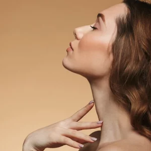 Profile view of a woman with long wavy hair, gently touching her neck while tilting her head back against a neutral beige background.