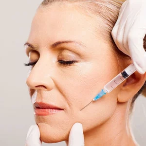 Mature woman receiving a cosmetic facial injection in the cheek area by a professional wearing white medical gloves, commonly associated with dermal filler or anti-wrinkle treatment.