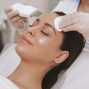 A woman receiving a facial treatment at a skincare clinic; a professional wearing gloves uses a cotton pad and a skin scrubber device on the woman's forehead while she relaxes with eyes closed.