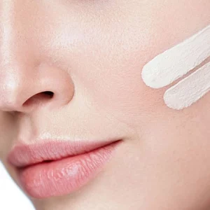 Close-up of a woman's face showing smooth skin and natural makeup, with two swipes of white cream or skincare product applied to her cheek, indicating a cosmetic or skincare application.