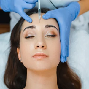Woman receiving a cosmetic forehead injection, likely Botox or dermal filler, administered by a professional wearing blue medical gloves. The patient is lying down with eyes closed during the procedure.