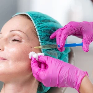 A woman wearing a surgical cap receives a cosmetic injection near her cheekbone from a medical professional in pink gloves, who is also holding a cotton pad to the skin.