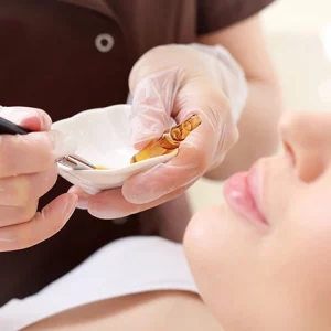 Esthetician wearing gloves preparing a facial treatment mixture in a small bowl with a brush and ampoule, next to a relaxed woman lying down for a skincare procedure.