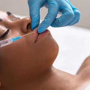 Woman receiving a cosmetic lip injection from a medical professional wearing blue gloves, lying down during the non-surgical aesthetic treatment.