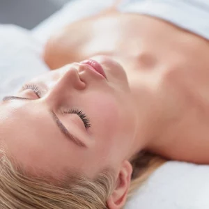A relaxed woman lying down with her eyes closed, receiving a facial or spa treatment, showcasing smooth, glowing skin and a calm atmosphere.