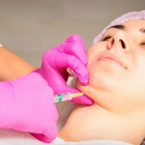Woman receiving a cosmetic injection in the chin area from a medical professional wearing pink gloves, suggesting a non-surgical aesthetic treatment such as dermal fillers or fat-dissolving injections.