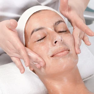 Woman with eyes closed receiving a facial treatment, as a professional gently massages a cream or cleanser into her skin, suggesting a soothing skincare or spa session.