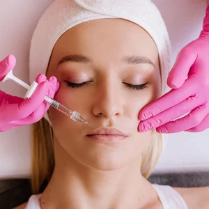 Woman lying down with eyes closed receiving a cosmetic injection in the cheek area by a practitioner wearing pink gloves, indicating a dermal filler or facial rejuvenation treatment.