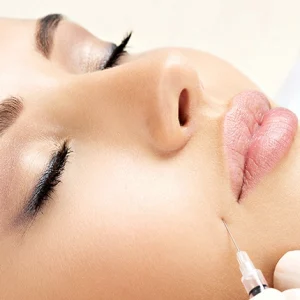 Close-up of a woman receiving a cosmetic injection in her lower cheek area while lying down with eyes closed, indicating a facial aesthetic treatment.