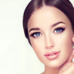 Close-up portrait of a woman with flawless skin, striking blue eyes, and elegant makeup, gently touching her face with one hand against a soft light background.