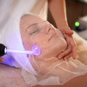 Woman receiving a high-frequency facial treatment with a violet electrode device applied over a gauze-covered face while lying relaxed on a spa bed. 24K Gold Facial Training Essex & London