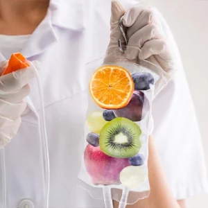 A healthcare professional wearing a white uniform and latex gloves holds an IV infusion bag filled with colorful fruits like kiwi, orange, apple, grapes, and blueberries, symbolizing a vitamin or nutrient IV therapy concept.