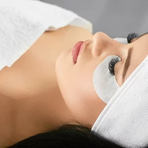 A woman lying down with her eyes closed, wearing a white headband and covered with a white towel. She has under-eye patches placed beneath her eyes, typically used for hydration or during eyelash extension procedures. Her eyelashes appear long and voluminous, indicating a beauty or spa treatment session.