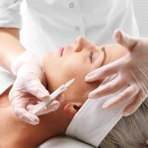 A woman lying down with eyes closed receiving a cosmetic injection in the temple area. A medical professional wearing white gloves is administering the injection and gently holding the patient's head for support.