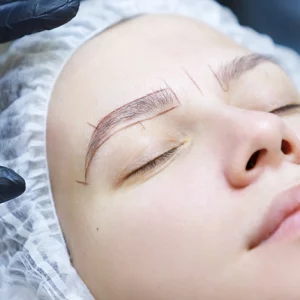Close-up of a woman preparing for an eyebrow microblading procedure, with guideline marks drawn on her eyebrows while wearing a disposable hair cap. A professional's gloved hand is positioned near her forehead.