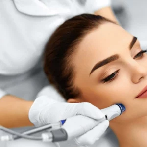A woman undergoing a professional facial treatment, receiving a hydrafacial or microdermabrasion procedure on her cheek using a specialized handheld device operated by a practitioner wearing white gloves.