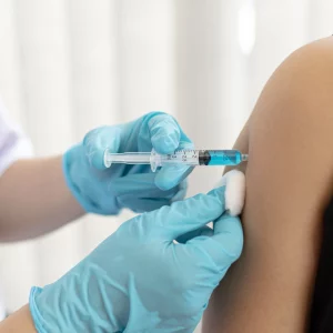 Healthcare professional wearing blue gloves administering an injection into a patient's upper arm using a syringe filled with blue liquid.