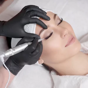 A woman lying down with her eyes closed while a beautician wearing black gloves performs a permanent eyeliner tattoo procedure using a handheld cosmetic tattoo machine.
