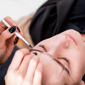 A beauty technician with black polished nails is carefully mapping out eyebrow shapes using a white pencil on a woman lying down with her eyes closed, in preparation for an eyebrow shaping or microblading procedure.