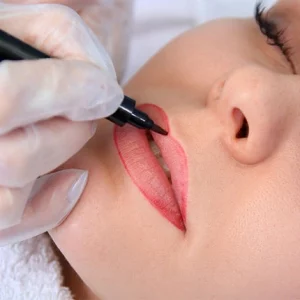A close-up of a woman lying down with her eyes closed while a technician wearing gloves outlines her lips with a cosmetic tattoo pen, preparing for a permanent lip makeup procedure.