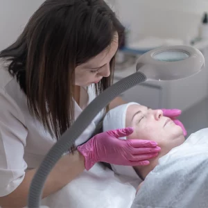 A female esthetician wearing pink gloves examines a woman's face under a magnifying lamp during a facial skincare treatment in a clinical or spa environment.