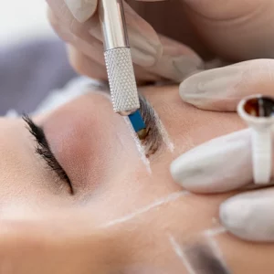 Close-up of a cosmetic procedure where a professional wearing gloves performs microblading on a woman's eyebrow using a handheld tool, with white guideline markings visible on the skin.