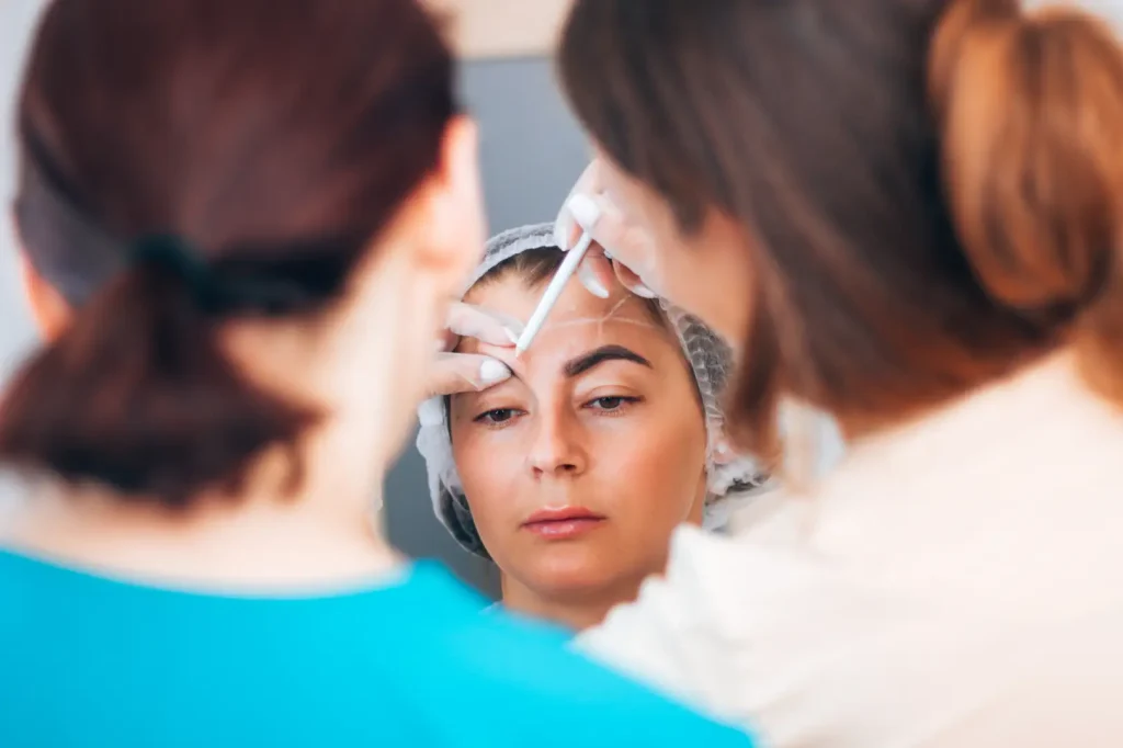 Two practitioners examine a woman's forehead while applying facial markings during an aesthetics training session.