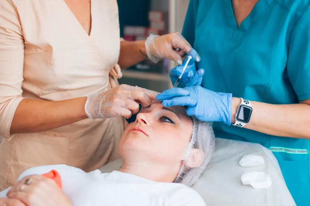 Two gloved practitioners work around a woman's forehead during a supervised injectable aesthetics procedure.