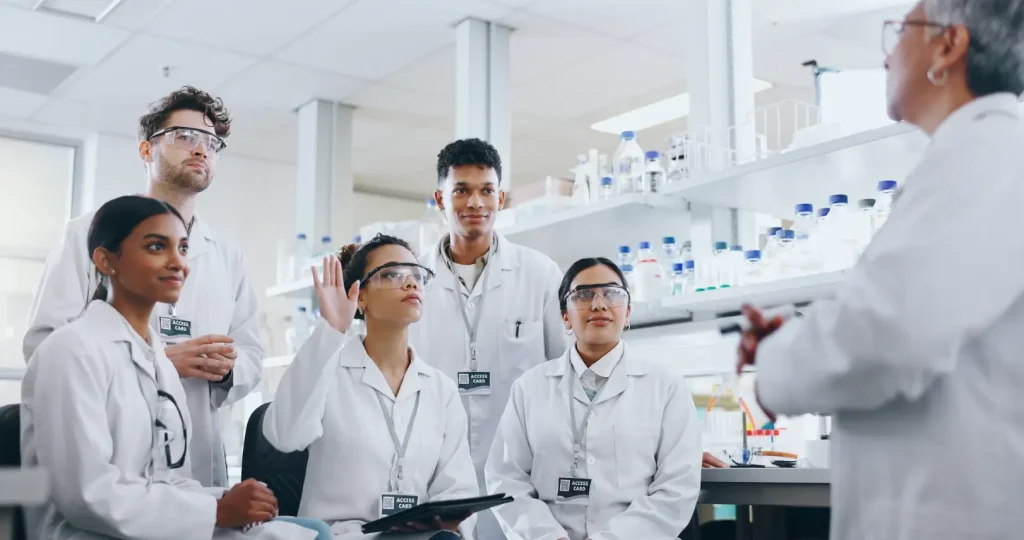 Group in white lab coats and safety glasses listening to an instructor in a laboratory.