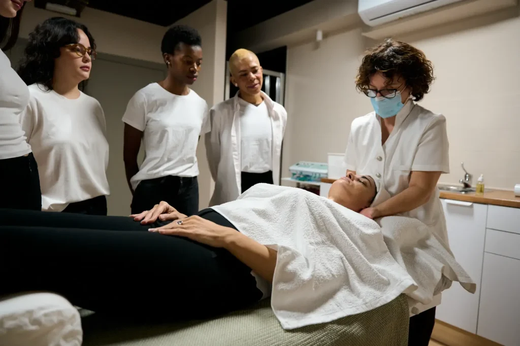 A practitioner demonstrates a facial technique on a reclining woman while several students observe.
