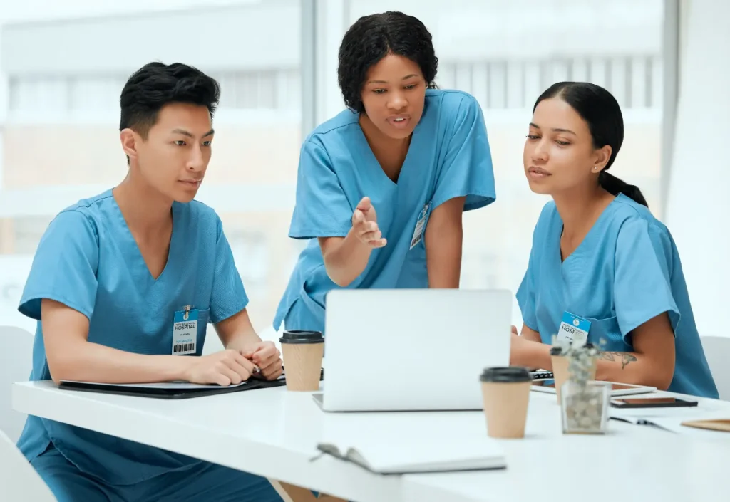 Three healthcare professionals in blue scrubs gather around a laptop during a group discussion.