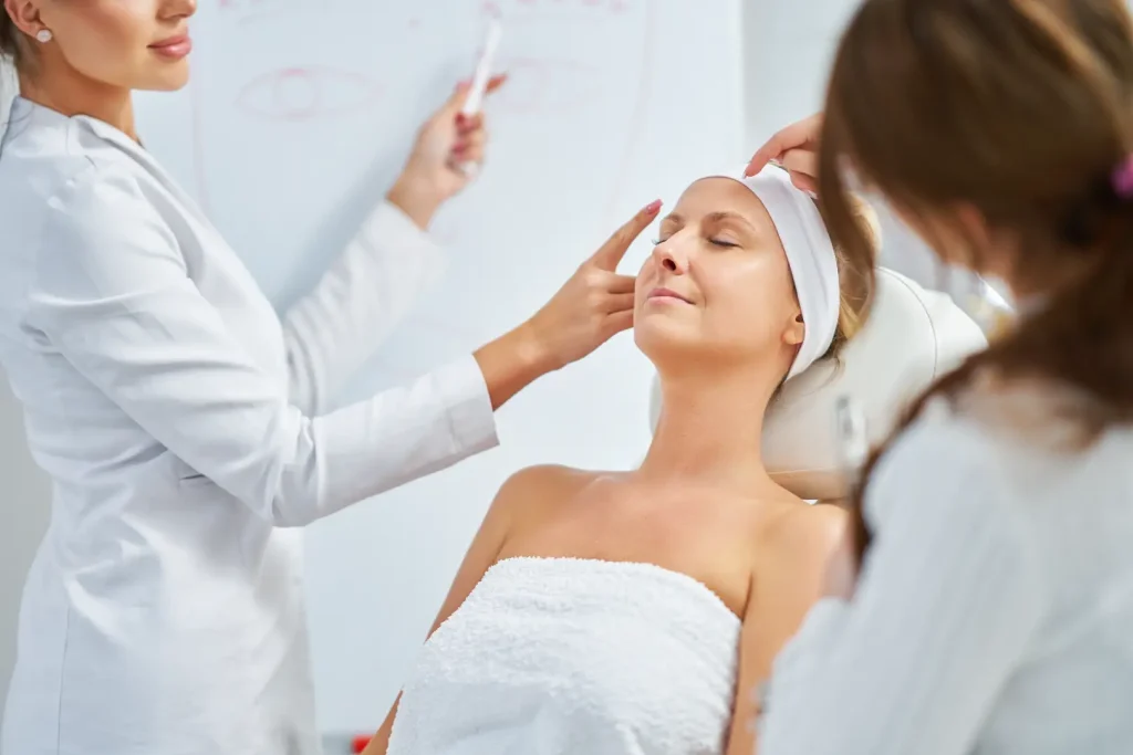 Two practitioners observe and demonstrate facial assessment techniques on a seated woman in a clinical setting.
