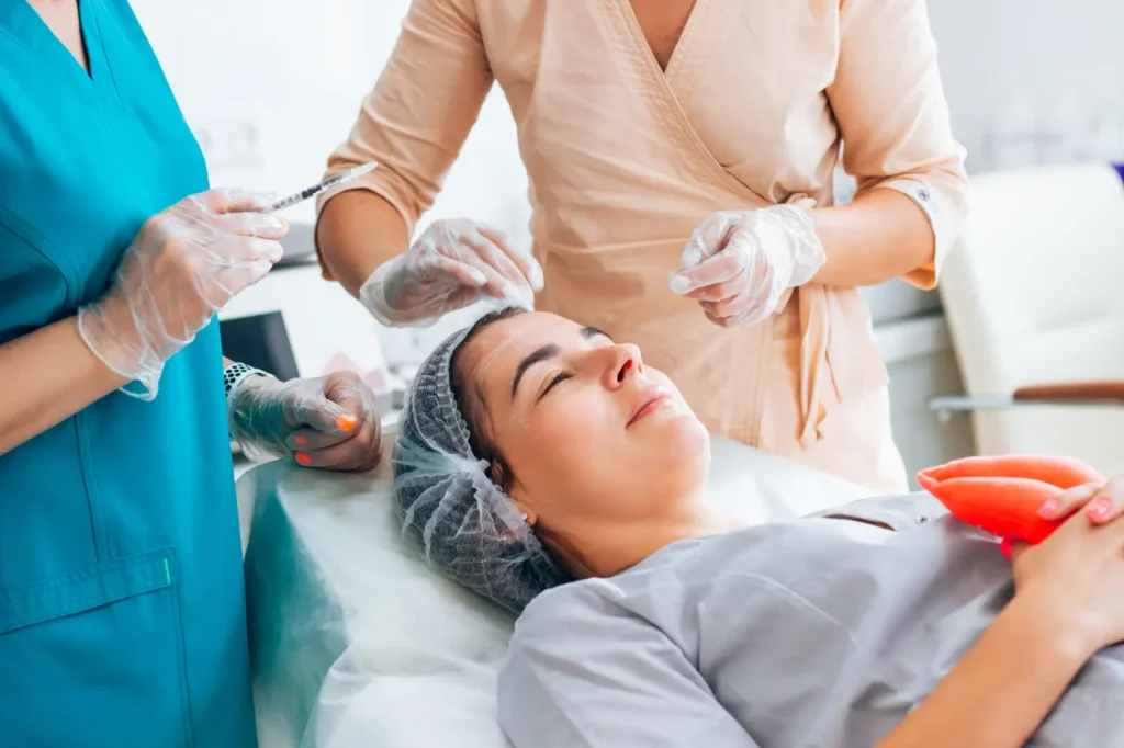 Two gloved practitioners work around a reclining woman's forehead while one holds a syringe.